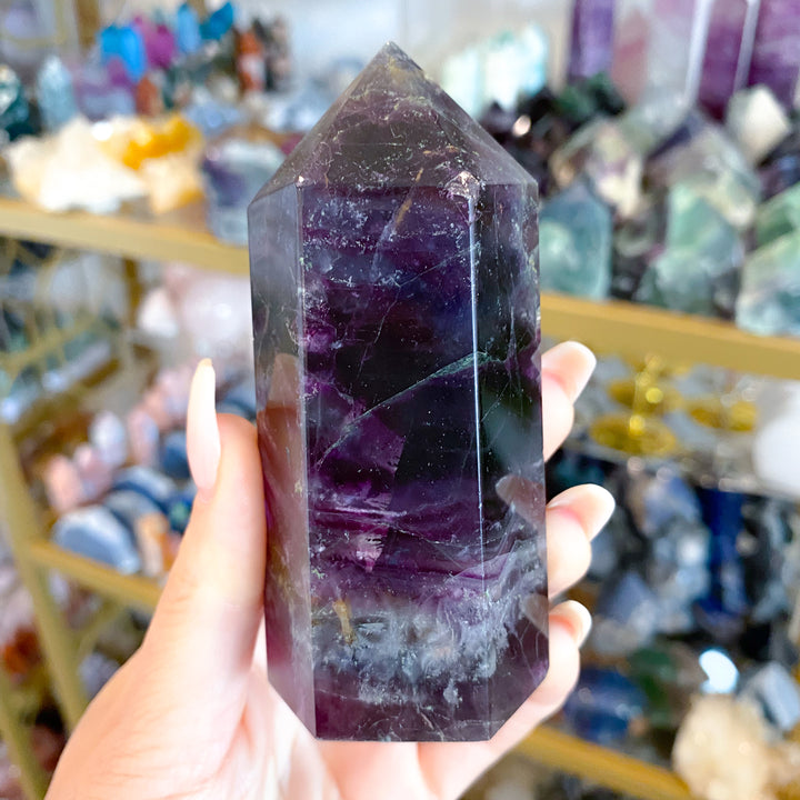 Hand holding a purple crystal tower in a store setting with various crystals in the background.