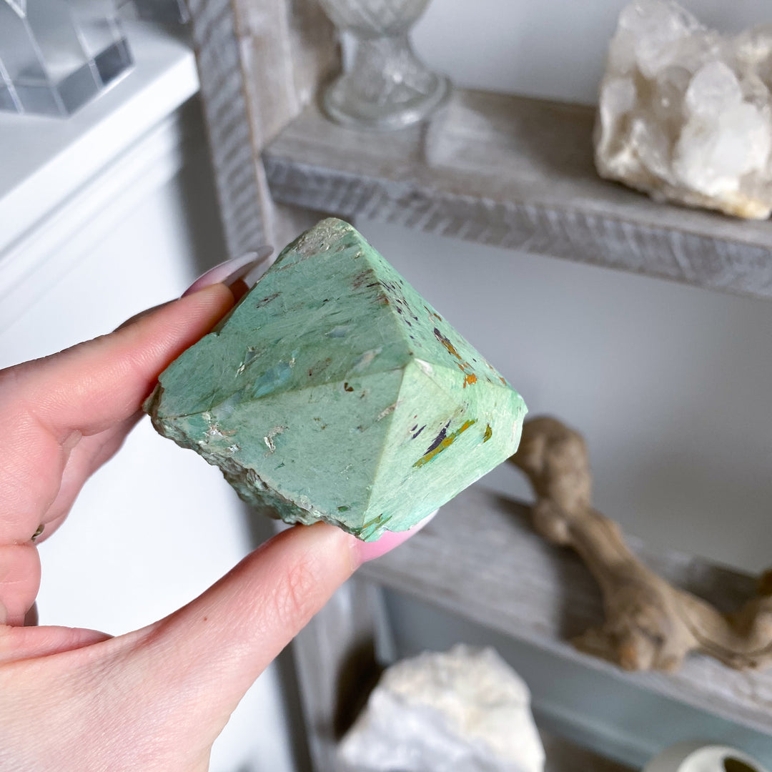 Hand holding a green crystal-like object with a blurred background of decorative items.