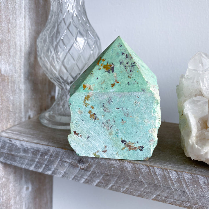 Turquoise crystal on a wooden shelf with a glass container and another crystal in the background.