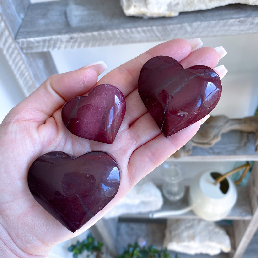 Hand holding three polished red stones shaped like hearts with a blurred indoor background.