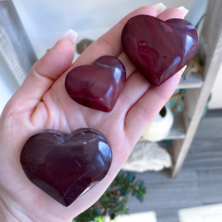 Three heart-shaped maroon stones held in a hand with a blurred indoor background