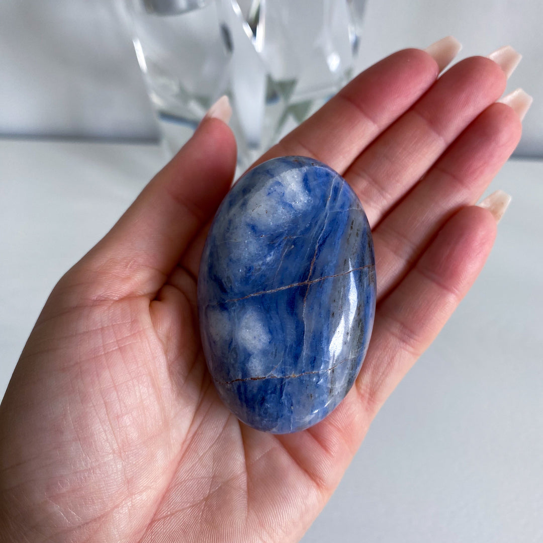 Hand holding a blue and white marbled stone with blurred glass objects in the background
