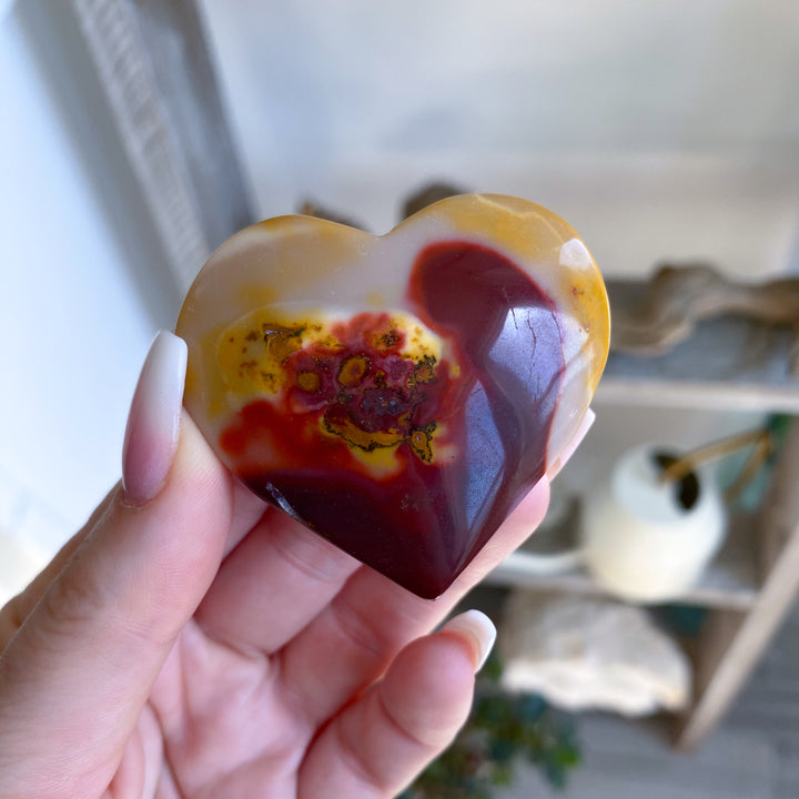 Heart-shaped stone with a colorful pattern held in a hand against a blurred indoor background